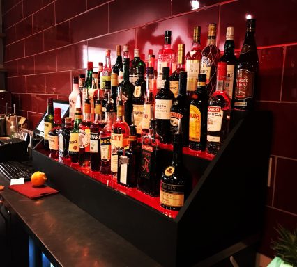 Display of various liquor bottles on a black shelf against a red tiled backdrop.