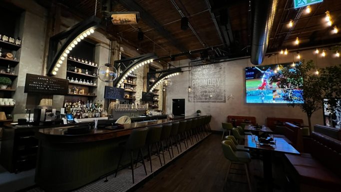 A modern café interior featuring a bar, seating area, and a large screen on the wall.