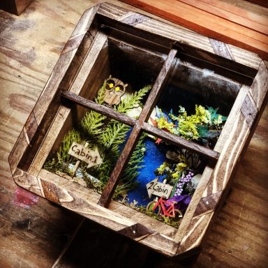 A wooden box divided into four sections, each featuring colorful miniature scenes with plants.